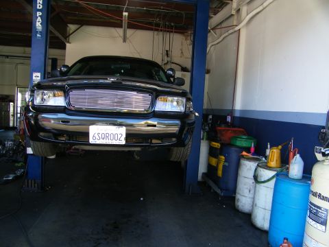 2003 Ford Crown Victoria - Front of black sedan raised on two-post lift, California plate 6SQR002, shop barrels at right