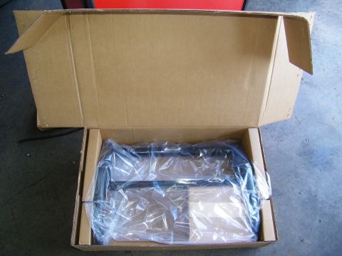 2003 Ford Crown Victoria - Plastic-wrapped black metal part inside an open shipping box on a garage floor
