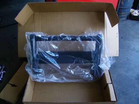 2003 Ford Crown Victoria - Open shipping box holding a plastic-wrapped black grille guard on the garage floor