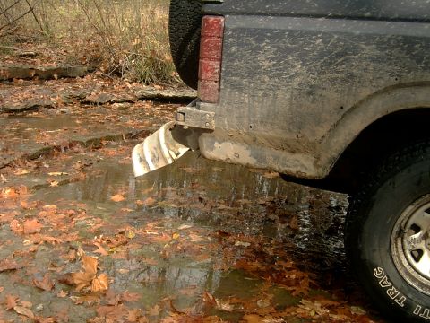 1982 Ford Bronco - This rear view of a muddy SUV shows its quarter panel and tire submerged in water with fallen leaves scattered around.