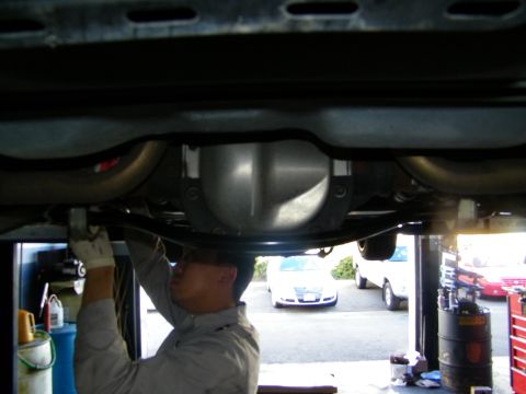 2003 Ford Crown Victoria - Technician reaching into the undercarriage at the differential, a sedan visible in the lot behind