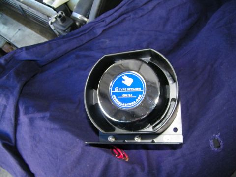 2003 Ford Crown Victoria - Chrome-faced HornBlasters 100W 8-ohm Omega speaker resting on dark blue fabric near vehicle parts