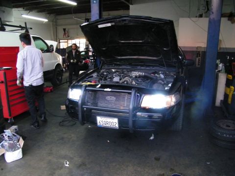 2003 Ford Crown Victoria - Black sedan with push-bar bumper guard, headlights on, hood open in repair shop bay