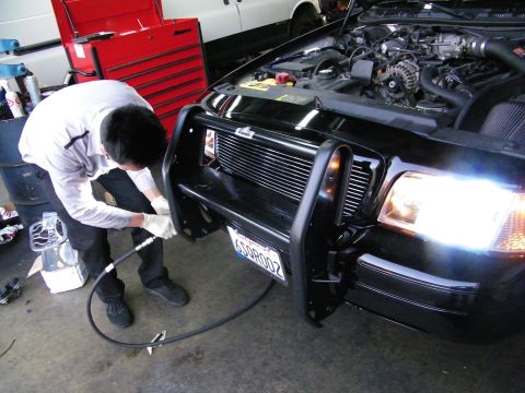 2003 Ford Crown Victoria - Mechanic using pneumatic gun at front bumper guard of black car, California plate visible