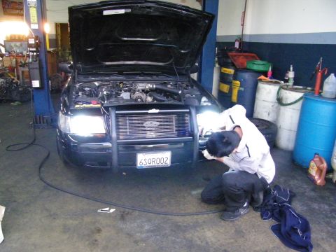 2003 Ford Crown Victoria - Worker at push-bar bumper of black car, hood open, oil drums and tires in background