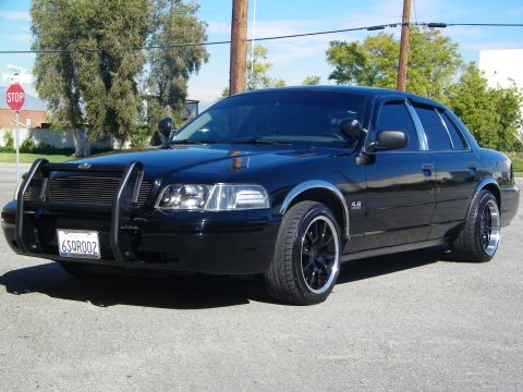 2003 Ford Crown Victoria - Front three-quarter of blacked-out sedan on dark wheels, 4.6 fender badge, trees behind