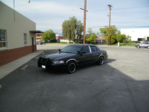 2003 Ford Crown Victoria - Black sedan with dark wheels and push bar sits at an intersection near a brick building.