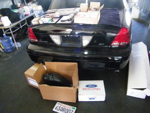 2003 Ford Crown Victoria - Black sedan trunk and taillights with Ford Racing performance parts boxes on the floor and a CA license plate.