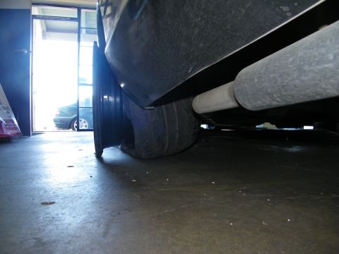2003 Ford Crown Victoria - Ground-level shot beneath a low black car showing tire, exhaust pipe and shop floor.