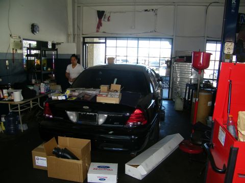 2003 Ford Crown Victoria - Black sedan parked in a service bay with a person nearby and Ford Racing boxes around it.