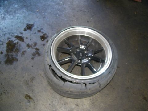 2003 Ford Crown Victoria - Polished-lip ten-spoke wheel with a split, delaminating tire lying on a damp concrete floor.