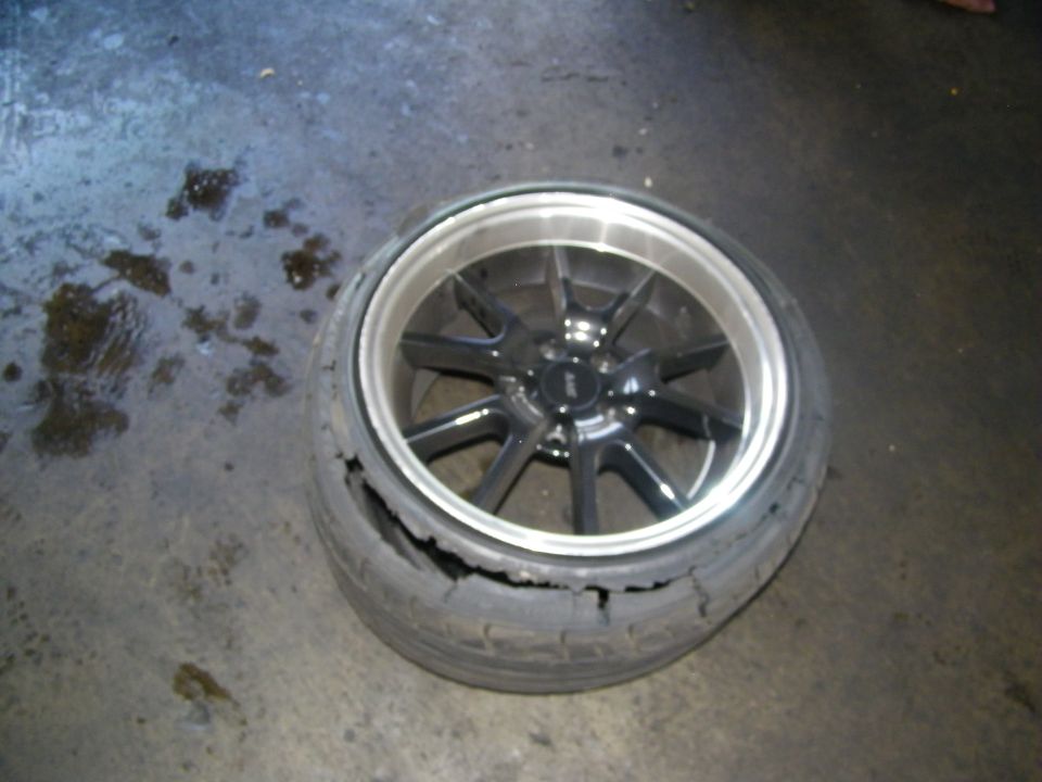 Gunmetal ten-spoke alloy wheel with cracked shredded tire on wet shop floor