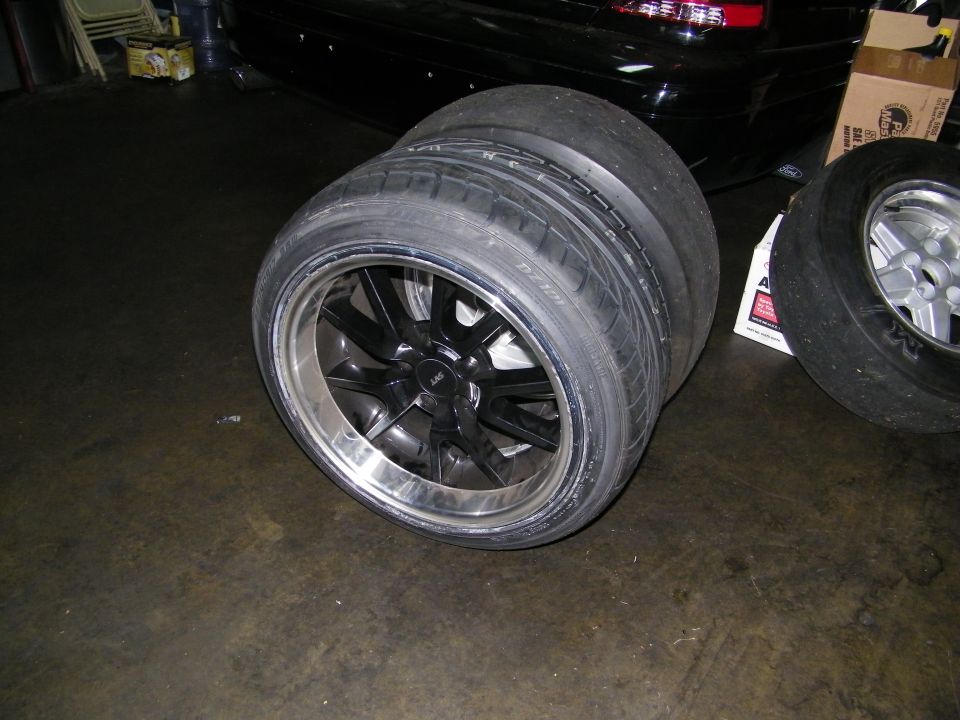 Two black-center polished alloy wheels with wide tires beside dark sedan in garage