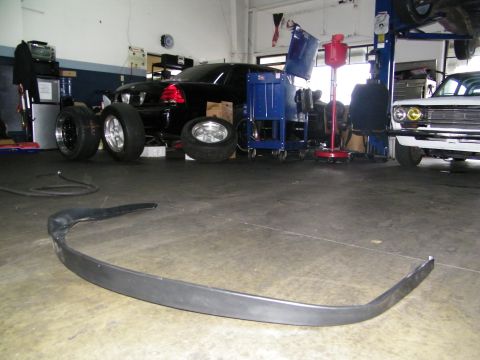 2003 Ford Crown Victoria - Unpainted steel front bumper on shop floor with a black sedan and classic car behind