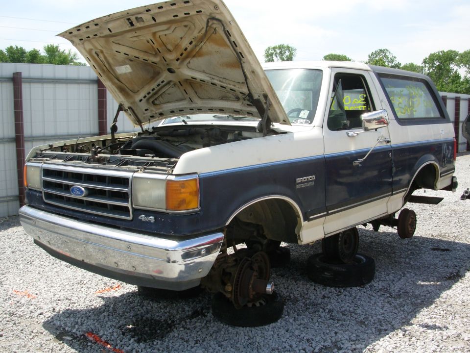 Blue and white Ford Bronco SUV with hood up in a junkyard