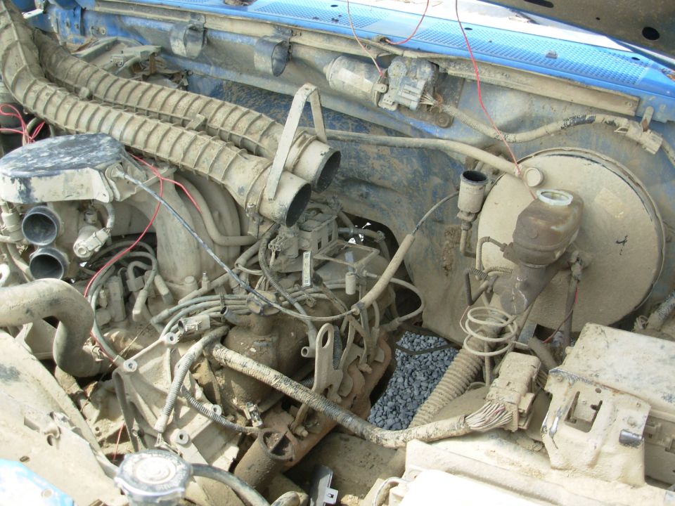 Dust-caked engine bay of a blue truck with coated wiring, air tube and carburetor