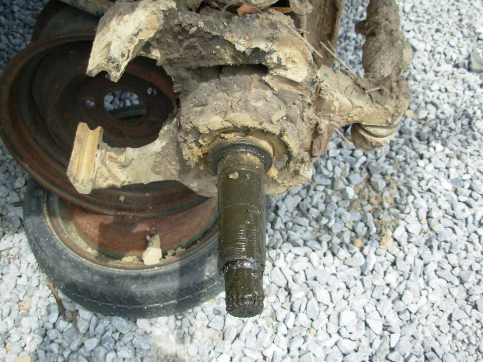 Mud-caked front axle knuckle and spindle on gravel beside a steel wheel