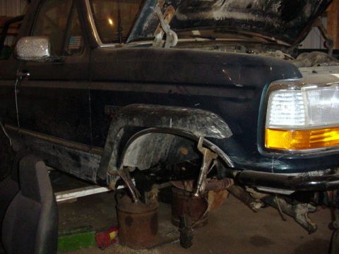 1992 Ford Bronco - Dark green Ford Bronco up on rusty buckets inside a shop with the hood open and the right front wheel, tire and suspension removed, exposin…