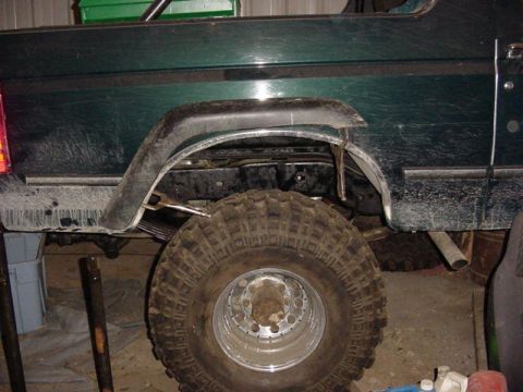 1992 Ford Bronco - Rear quarter of a dark green lifted truck body with the wheel opening cut and trimmed for clearance, exposing bare metal at the arch edge a…