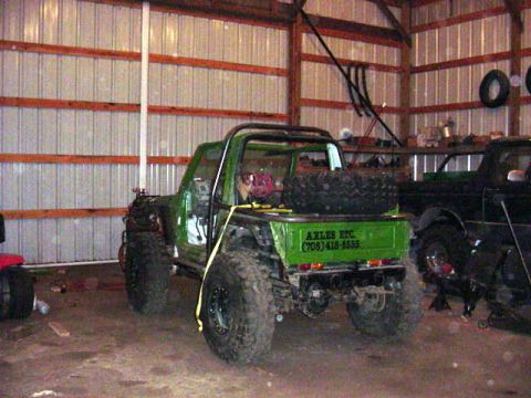 1992 Ford Bronco - A green off-road vehicle is parked in a garage next to a black truck, with a spare tire mounted in the bed.