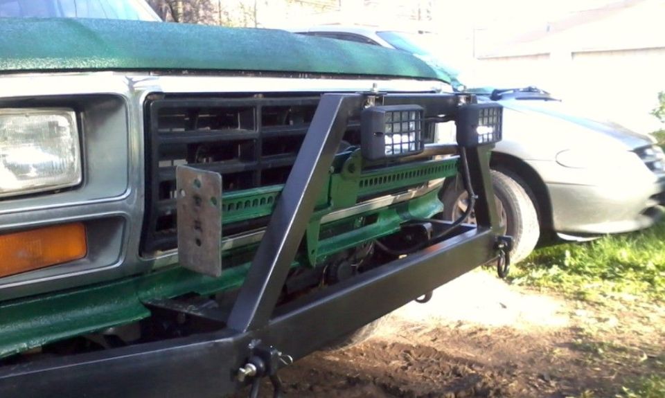 Green truck front end with custom black bumper, Hi-Lift jack, and two square lights. Another car is visible to the right