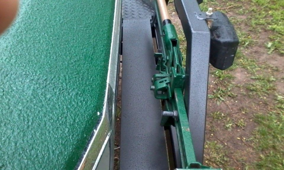 Green latch linkage mechanism on gray metal frame next to textured green panel, grass in background