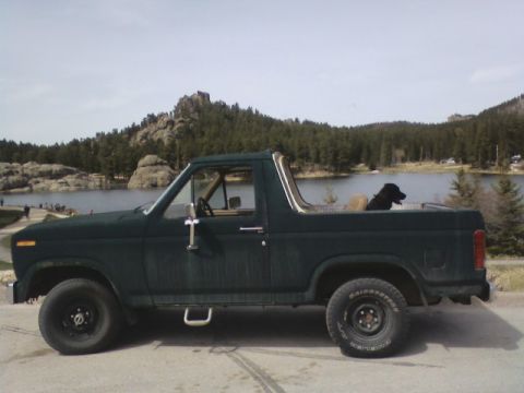 1986 Ford Bronco - This SUV is parked near a lake with a scenic view of hills and trees under a cloudy sky.