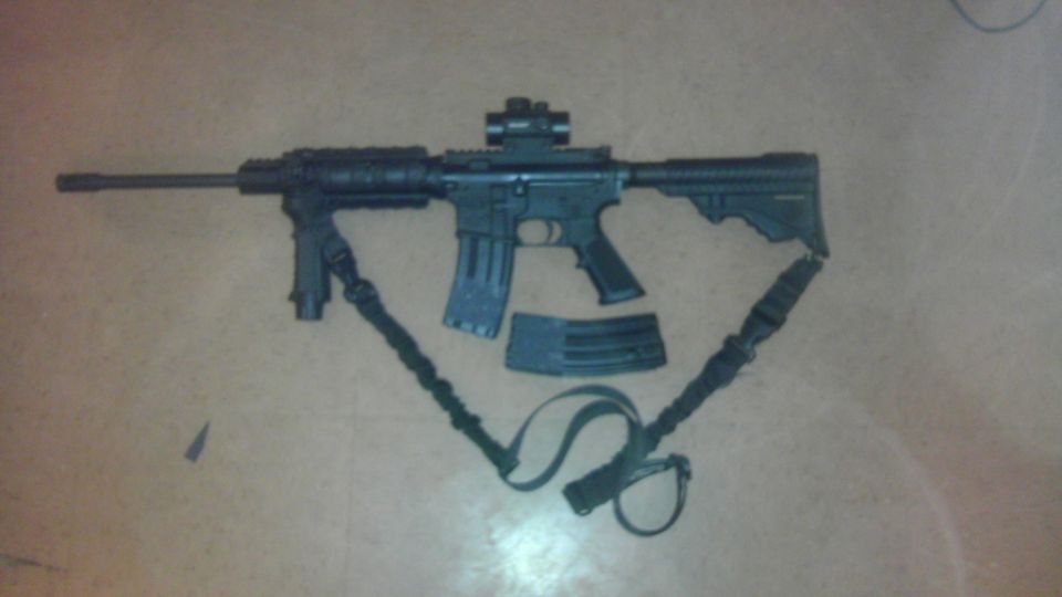 AR-15 style rifle with a red dot sight, foregrip, and two magazines. A sling is attached