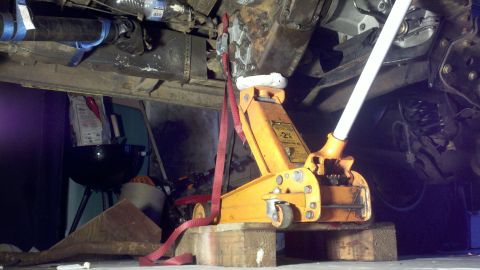 1978 Ford Bronco - The underside of a vehicle is shown, lifted by a jack and secured with a strap, with various mechanical parts visible.
