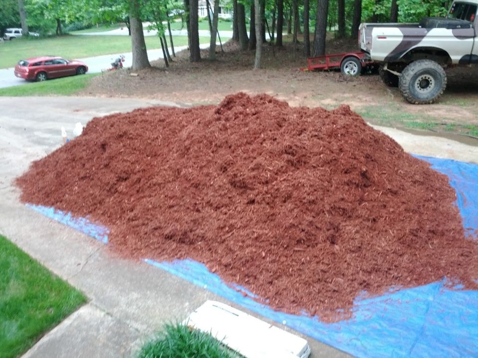 Pile of red mulch on a blue tarp with a red car and a pickup truck in the background
