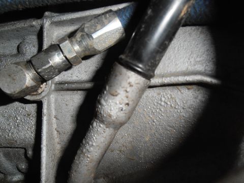 1990 Ford Bronco - Dark under-hood close-up of the side of a cast aluminum transmission case, showing a metal dipstick tube pressed into its bore alongside a…
