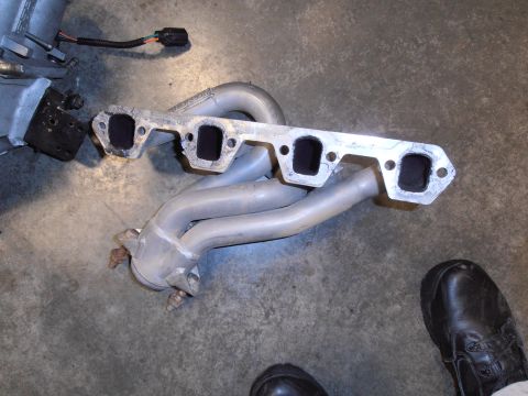 1990 Ford Bronco - Exhaust manifold part shown on concrete floor, with a shoe visible in the lower right corner.