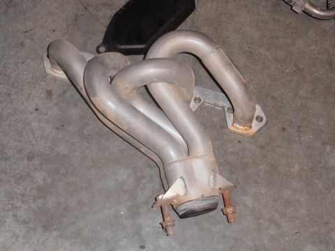 1990 Ford Bronco - A silver exhaust manifold with visible bolts and mounting plates is shown on a concrete surface.