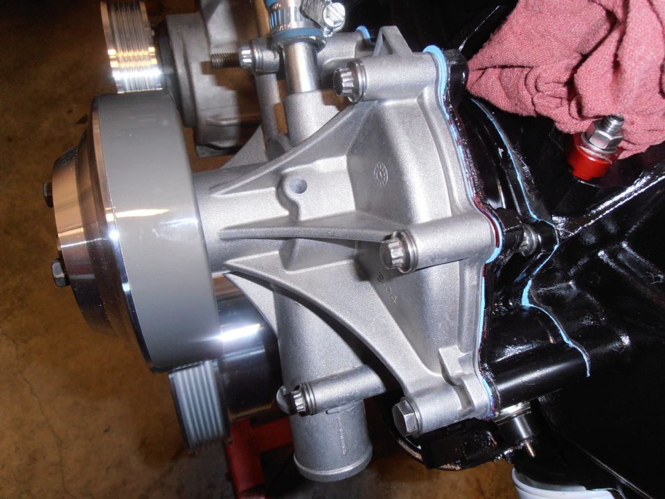 Silver aluminum engine water pump with blue gasket sealant attached to a black engine block