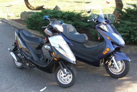 2008 Yamaha Zuma - Two scooters are parked outdoors on a paved surface near some bushes and trees.