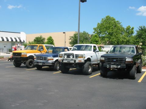 1992 Ford Bronco - Four lifted trucks are parked in a lot on a sunny day.