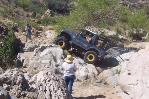 1981 Ford F-150 - A dark blue off-road vehicle navigates a challenging rocky trail with people observing.
