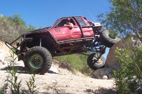 1981 Ford F-150 - A heavily modified red truck with a roll cage is shown off-roading on a rocky terrain.