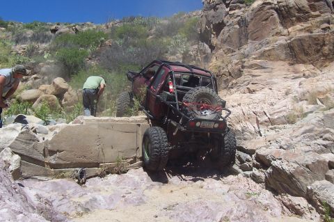 1981 Ford F-150 - A modified pickup truck is being driven over a rough, rocky obstacle course with people watching.
