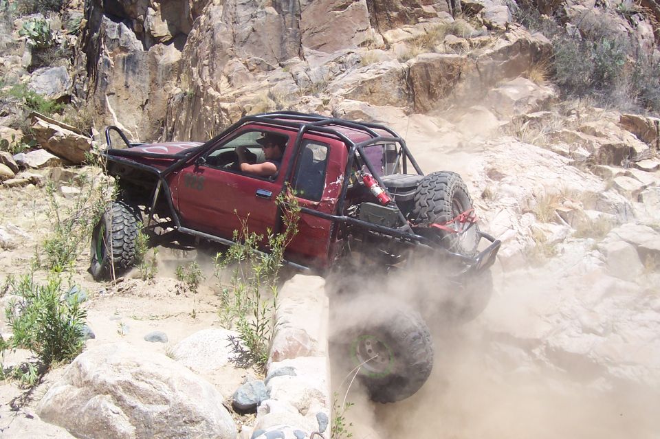 Red off-road truck with a roll cage and spare tire navigating a rocky, dusty trail