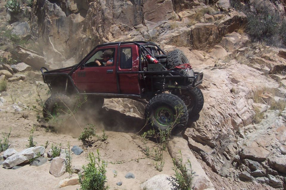 Modified red pickup truck with roll cage and large tires climbing a dusty, rocky hill