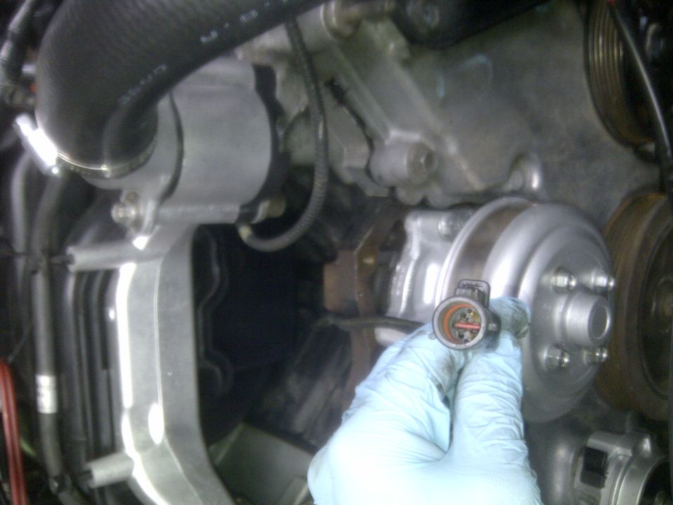 Close-up of a hand in a blue glove holding a circular electrical connector with orange and red wires, near a silver engine component