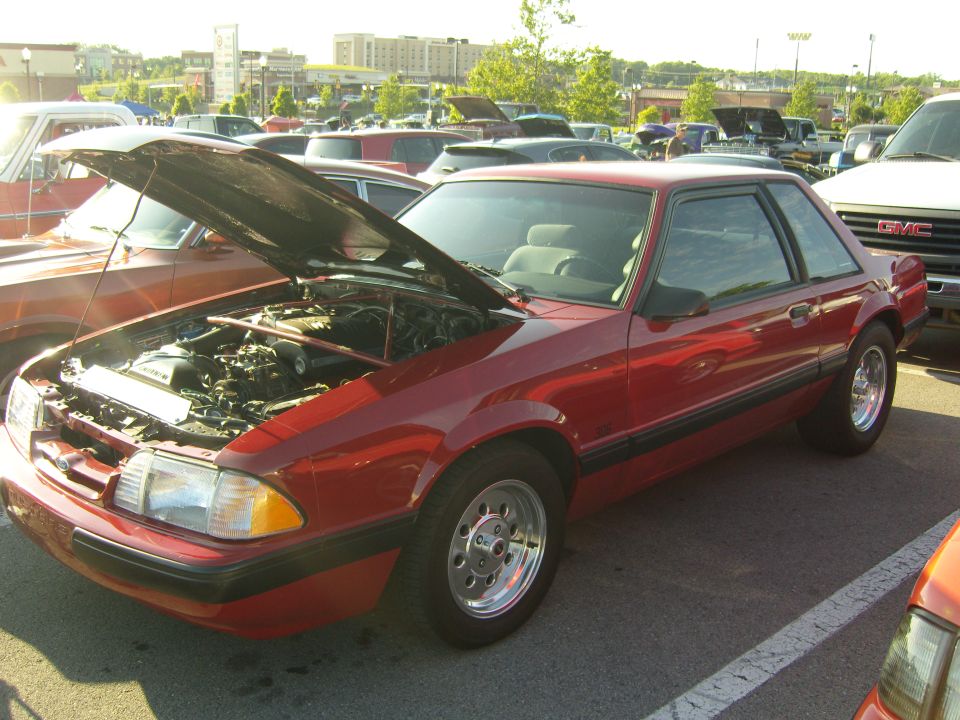 Red coupe with hood open, showing engine bay. Other cars parked nearby