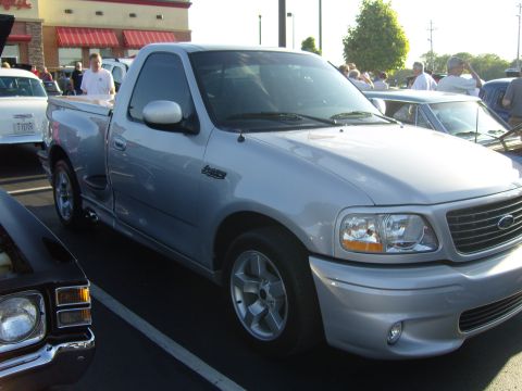 2007 Ford Mustang - A silver Ford F-150 Lightning pickup truck is parked at an outdoor event with other vehicles and people visible.