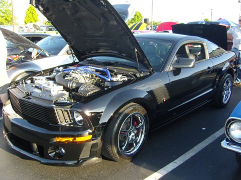 2007 Ford Mustang - Black 2007 Ford Mustang with hood open, displaying its supercharged engine at a car show.