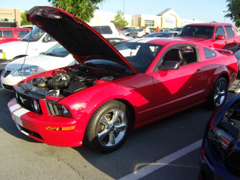 2007 Ford Mustang - A red Ford Mustang coupe with its hood open, parked in a lot with other vehicles and a building in the background.
