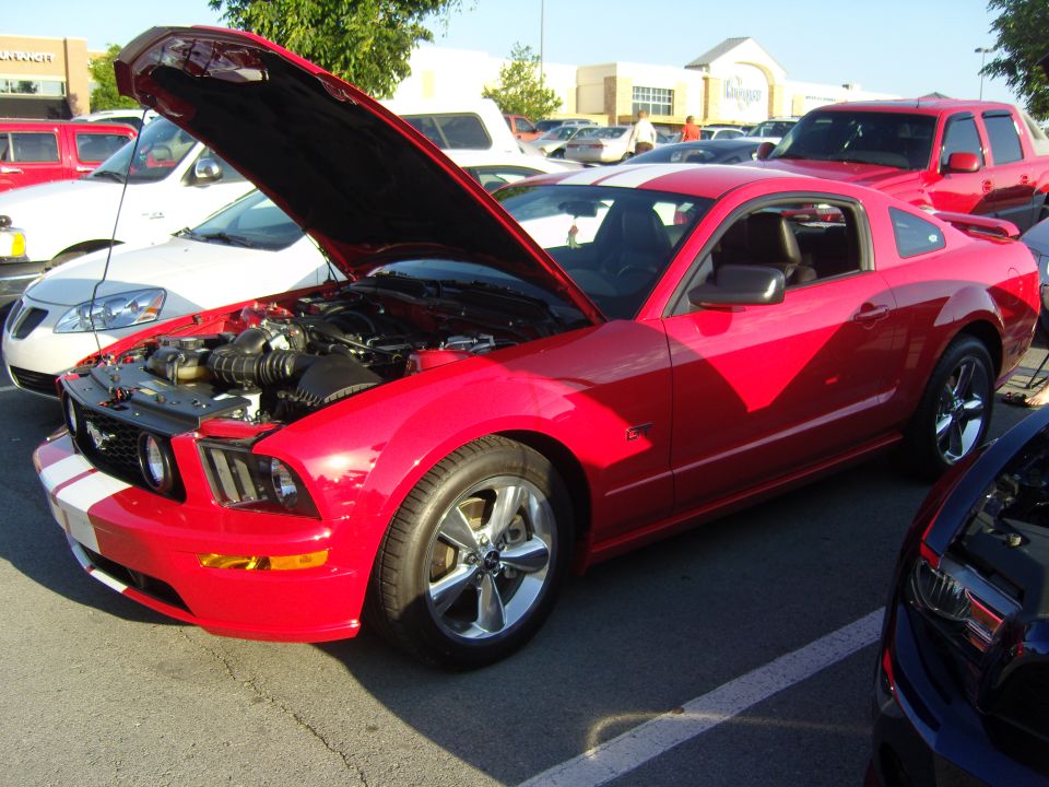 Red Ford Mustang coupe with white racing stripes, hood open revealing engine, parked in a lot