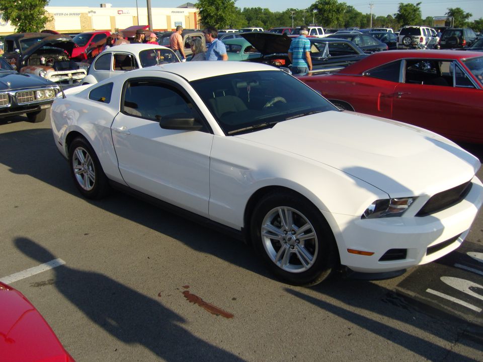 White 2010 Ford Mustang coupe parked on asphalt, front passenger side visible, with red car in foreground and other vehicles in background