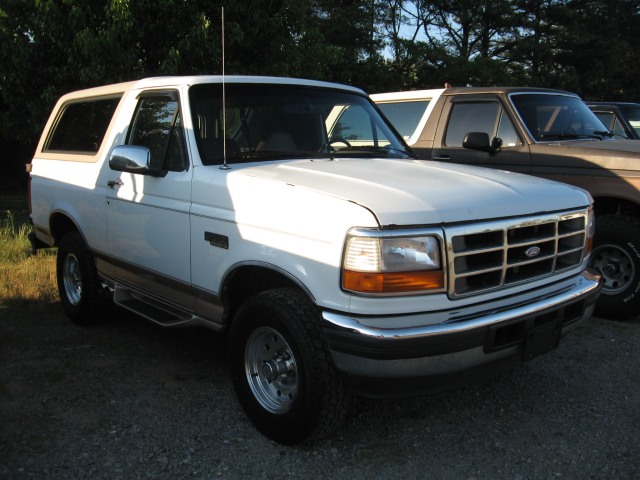 White Ford Bronco from the front-right, showing chrome grille and side steps. Another Bronco is partially visible behind it