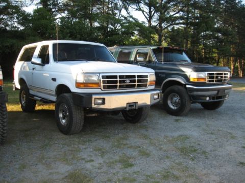 1978 Ford Bronco - Two Ford Bronco SUVs are parked side-by-side on a gravel lot with trees in the background.